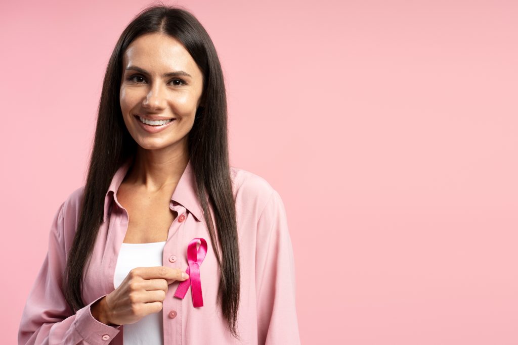 Portrait of smiling woman showing pink ribbon attached to her shirt, symbolizing breast cancer awareness, against a pink background, promoting health and hope. Health care concept